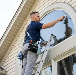 Professional window cleaner on a ladder using a squeegee to clean an exterior arched window, Eugene, OR.